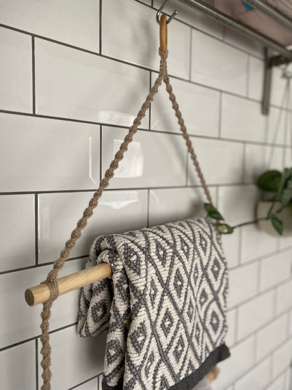 Close up of a bathroom towel holder, made with taupe yarn, wooden dowels and a wooden ring, hanging against a white tiled wall, holding a grey and white patterned towel. 